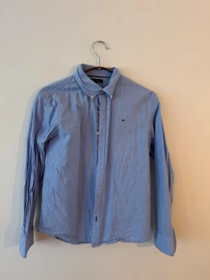 Blå skjorta med knapparTommy Hilfiger - Klassisk blå skjorta med lång ärm och knappar framtill. Skjortan har button down-krage och en liten broderad logga på bröstet. Perfekt till både jeans och chinos för en clean look.