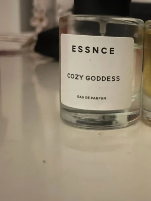 Essnce Cozy Goddess Eau de Parfum - Essnce Cozy Goddess Eau de Parfum i en rund, genomskinlig glasflaska med svart lock. Doften presenteras i en stilren design med vit etikett och svart text. Perfekt för dig som gillar minimalistiska och moderna parfymflaskor.