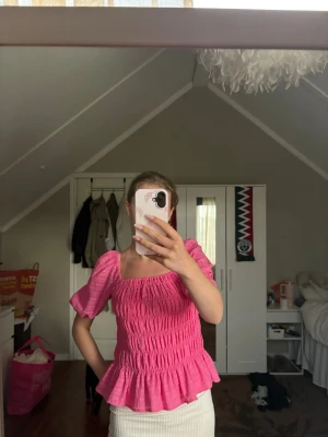 Rosa smockad topp med puffärm - Trendig rosa topp med smockad kropp och volang nedtill. Toppen har puffiga korta ärmar och fyrkantig ringning både fram och bak. Perfekt för dig som vill ha en söt och färgglad look.