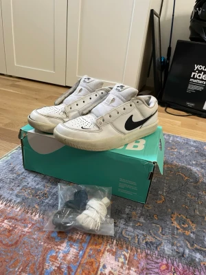 Nike SB Dunk Low vita sneakers - Nike SB Dunk Low sneakers i vitt läder med svart swoosh på sidan. Klassisk låg siluett med perforerad tåbox och vadderad plös. Kommer med extra skosnören i svart och vitt. Perfekta för dig som gillar streetwear och skate.