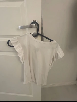 Vit topp med volangärmar - Söt vit topp med korta volangärmar och rund halsringning. Perfekt för en fräsch och enkel look. Tunn och mjuk i materialet, passar bra till både jeans och kjol.