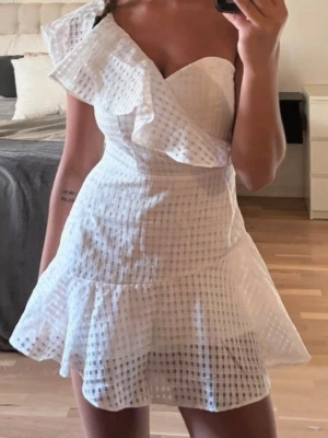 Vit omlottklänning med volang - Snygg vit kortklänning med rutigt mönster och volangdetalj över ena axeln. Klänningen är axelbandslös på ena sidan och har en omlott-look upptill. Perfekt för sommarens alla tillfällen.