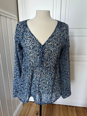 Blommig blå transparent blus - Snygg blus i transparent blått tyg med små vita och ljusblå blommor. Djup v-ringning och knyt framtill ger en avslappnad look. Långärmad modell i lätt och luftig polyester, perfekt för lager på lager. Passar till både jeans och kjol.