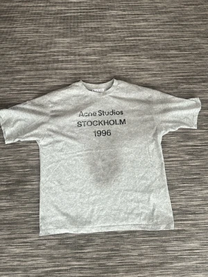 Grå Acne Studios t-shirt med tryck - Snygg grå t-shirt från Acne Studios med svart texttryck 'STOCKHOLM 1996' på bröstet. Klassisk passform med rund hals och korta ärmar. Perfekt för en stilren och avslappnad look.