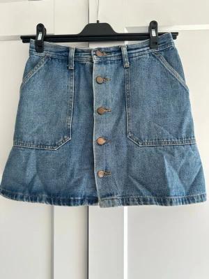 Blå jeanskjol med knappar - Snygg blå jeanskjol från My Style med framfickor och knappar längs hela framsidan. Kjolen har bälteshällor och klassisk denimlook, perfekt för dig som gillar en avslappnad och trendig stil.