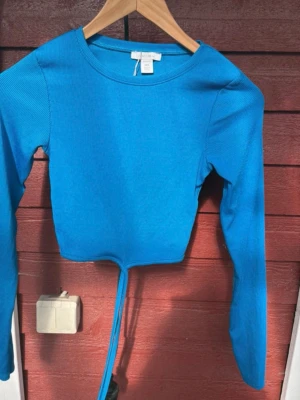 Blå ribbad croppad topp från Amisu - Snygg blå croppad topp från Amisu med långa ärmar och ribbad struktur. Toppen har en cool snörning framtill som ger en trendig look. Perfekt att styla med höga jeans eller kjol för en fräsch outfit.