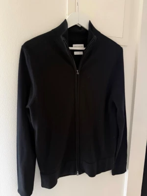 Svart ziptröja från These Glory Days - Svart ziptröja från These Glory Days i mjukt material. Tröjan har hög krage, hel dragkedja och lång ärm. Enkel och stilren design som passar perfekt till lager-på-lager. Materialet är merinoull vilket gör den extra skön.