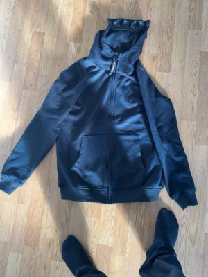 Svart zip-hoodie med fickor - Säljer en svart hoodie med dragkedja och stora framfickor. Tröjan har huva med metallöljetter och ribbade muddar vid ärmslut och nederkant. Perfekt för en chill och avslappnad stil. Materialet känns mjukt och bekvämt, troligen bomullsblandning.
