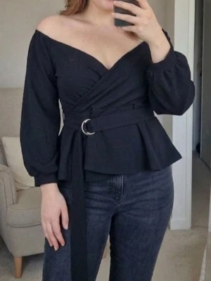 SÅLD Svart omlottopp med bälte - Trendig svart omlottopp med v-ringning och offshoulder-design. Toppen har långa ärmar, markerad midja med bälte och lätt utsvängd nederdel. Perfekt för en stilren look och enkel att matcha med jeans eller kjol. Storlek 44. Använd en gång. 