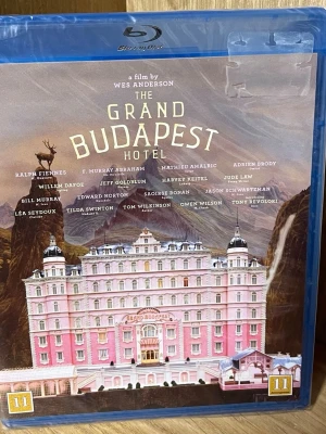 The Grand Budapest Hotel Blu-ray - Säljer en Blu-ray av filmen The Grand Budapest Hotel, regisserad av Wes Anderson. Omslaget är rosa med en bild på hotellet och listar skådespelare som Ralph Fiennes och Saoirse Ronan. Filmen är på engelska med svensk text och har åldersgräns 11 år.