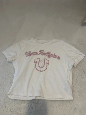 Vit True Religion t-shirt med tryck - Vit t-shirt från True Religion med rosa text och logga framtill. Klassisk passform med korta ärmar och rund halsringning. Perfekt för en avslappnad och trendig stil.