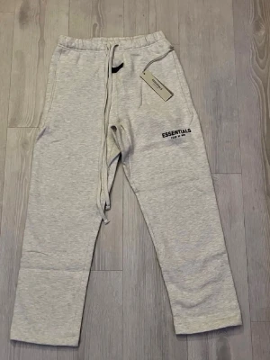 Grå hoodie från Essentials Fear of God - Snygg ljusgrå hoodie från Essentials Fear of God med svart logga tryckt på ryggen. Hoodien har huva, ribbade muddar och en avslappnad passform. Perfekt för en streetwear-look och tillverkad i mjukt bomullsmaterial.