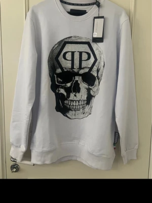 Vit Philipp Plein sweatshirt med dödskalle - Säljer en vit sweatshirt från Philipp Plein med ett stort svart dödskalletryck och PP-logga på bröstet. Tröjan har rund hals, ribbade muddar och en relaxed fit. Perfekt för dig som gillar streetwear och statementplagg.