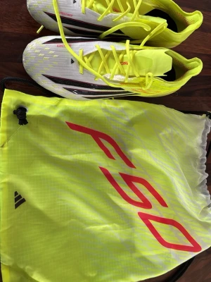 Adidas X Speedflow fotbollsskor gul/vit - Säljer ett par Adidas X Speedflow fotbollsskor i vitt och neon-gult med svarta detaljer. Skorna har snörning och låg profil, tillverkade i lätt syntetmaterial för maxad speed. Medföljer matchande gympåse i neon-gult med röd logga.