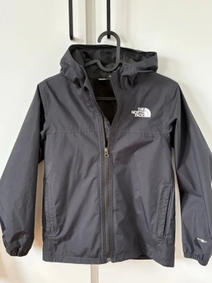Svart vindjacka från The North Face - Svart vindjacka från The North Face med huva och dragkedja framtill. Jackan har två sidofickor med dragkedja, elastiska muddar och logga på bröstet. Perfekt för blåsiga dagar och enkel att matcha med olika outfits. Jackan är i väldigt fint skick förutom att det är ett brännmärke längst ner på ryggen som knappt syns.