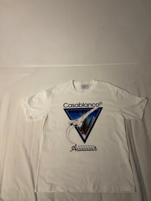 Vit Casablanca t-shirt med tryck - Vit t-shirt från Casablanca med stort grafiskt tryck framtill som visar ett flygplan och stadssilhuett i blått och svart. T-shirten har rund hals och korta ärmar. Perfekt för dig som gillar unika prints och streetwear vibes.