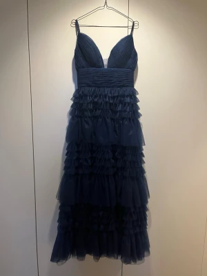 Marinblå balklänning med volanger - Snygg marinblå långklänning med smala axelband och djup v-ringning. Klänningen har flera lager av volanger i tyll och ett markerat midjeband. Materialet skimrar lätt och ger en elegant look. Perfekt för speciella tillfällen.