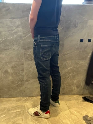 Mörkblå slim fit jeans från Replay - Snygga mörkblå slim fit jeans från Replay med klassiska fem fickor och diskreta slitningar. Jeansen har vita kontrastsömmar och en modern passform som sitter tajt längs benen. Perfekta för en trendig och avslappnad stil.