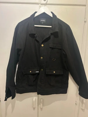 Vårjacka - Svart jacka från Theories med flera fickor framtill och guldiga knappar. Klassisk krage och rak passform, tillverkad i bomull. Perfekt för dig som gillar streetwear och enkel stil.
