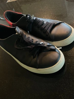 Svarta sneakers från argato - Stilrena svarta sneakers från arigato med vita sulor och röda detaljer vid hälen. Skorna har svarta skosnören och är tillverkade i slätt lädermaterial. Perfekta för en clean och enkel look.