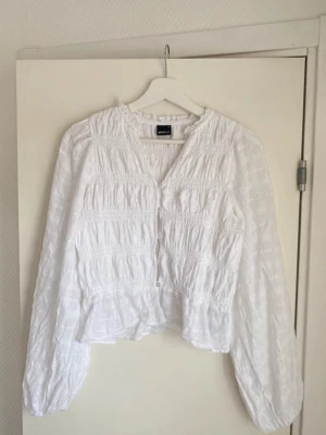 Vit blus med knappar från Gina Tricot - Söt vit blus från Gina Tricot med lång ärm och knappar framtill. Blusen har smockdetaljer över bröstet och lätt puffiga ärmar. Tillverkad i ett tunt, luftigt tyg med diskret rutmönster. Perfekt för en trendig och avslappnad look.