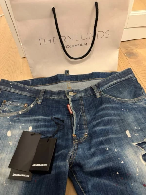 Disqaured jeans - Snygga blå jeans från DSQUARED2 med coola färgstänk och slitningar. Klassisk femficksmodell med gylf och knapp, samt detaljer i gult och rött. Jeansen är tillverkade i bomull och har en trendig, avslappnad look.
