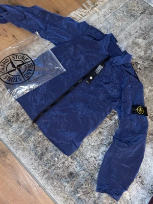 Blå vindjacka från Stone Island - Snygg blå vindjacka från Stone Island med ikonisk patch på vänster ärm. Jackan har hel dragkedja framtill, ståkrage och är tillverkad i lätt, glansigt material som ger en modern look. Perfekt för dig som vill ha en stilren och sportig jacka.