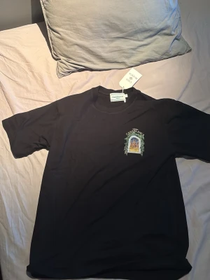 Svart Casablanca t-shirt med tryck - Säljer en svart t-shirt från Casablanca med färgglatt tryck på bröstet och stort grafiskt tryck på ryggen. T-shirten har rund hals och korta ärmar. Perfekt för dig som gillar streetwear och vill sticka ut med snygg design.