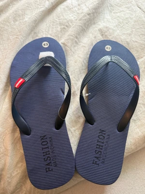 Mörkblå tofflor - Mörkblå flip-flops/tofflor från Fashion med svarta remmar och små röda detaljer vid tårna. Sulan är ribbad för extra grepp och komfort. Perfekta för stranden eller sommardagar. Tillverkade i syntetmaterial. Aldrig använda, fått i present helt nya. 