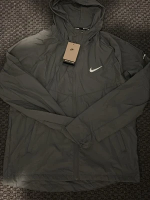 Svart Nike vindjacka med huva - Snygg svart vindjacka från Nike med vit logga på bröstet. Jackan har huva, dragkedja framtill och två sidofickor. Tillverkad i lätt och tåligt material som passar perfekt för blåsiga dagar. Enkel och stilren design.