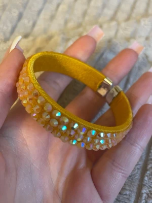 Gult armband med glittrande pärlor - Snyggt gult armband dekorerat med skimrande, multifärgade pärlor som reflekterar ljuset. Armbandet har en bred form och stängs med ett metallspänne. Perfekt accessoar för dig som vill sticka ut med färg och glitter.