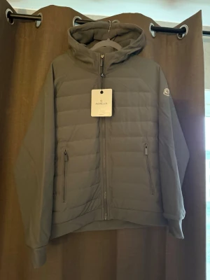 Grå dunjacka med huva från Moncler - Snygg grå dunjacka från Moncler med huva och två dragkedjeförsedda fickor framtill. Jackan har quiltad design och diskret Moncler-logga på ärmen. Perfekt för kalla vinterdagar och ger en stilren look.