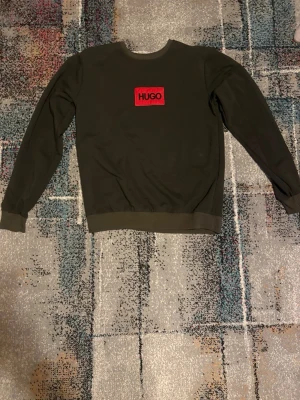 Mörkgrön sweatshirt från Hugo - Snygg mörkgrön sweatshirt från Hugo med röd broderad logotyp på bröstet. Tröjan har rund halsringning, långa ärmar och ribbade muddar vid ärmslut och nederkant. Perfekt för en avslappnad och trendig stil.