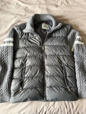 Grå pufferjacka från Moncler - Snygg grå pufferjacka från Moncler med stickade ärmar och vita ränder på ärmarna. Jackan har två dragkedjeförsedda fickor framtill, hög krage och Moncler-logga på vänster ärm. Kroppen är quiltad och vadderad medan ärmarna är kabelstickade.