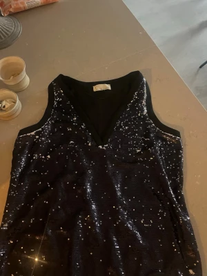 Svart paljettopp med v-ringning - Glittrig svart topp med v-ringning och ärmlös design. Toppen är täckt av paljetter som ger en festlig look och har en snygg passform. Perfekt för dig som vill sticka ut och glänsa.