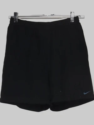 Svarta träningsshorts från Nike - Svarta träningsshorts från Nike med klassisk design och liten blå Nike-logga på benet. Shortsen har elastisk midja och är tillverkade i ett lätt och funktionellt material som passar perfekt för sport och aktivitet.