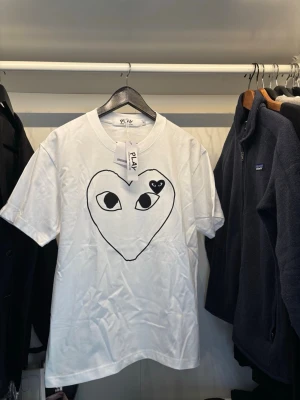 Vit t-shirt från Comme des Garçons Play - Vit t-shirt från Comme des Garçons Play med ett stort hjärta och ögon tryckt i svart på bröstet. Klassisk rund hals och korta ärmar. Perfekt för dig som gillar streetstyle och vill sticka ut med en ikonisk design.