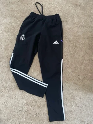 Svarta Adidas Real Madrid träningsbyxor - Svarta träningsbyxor från Adidas med Real Madrid-logga på ena benet och Adidas-logga på det andra. Byxorna har vita ränder längs sidorna och resår i midjan med dragsko. Perfekta för fotbollsträning eller chill.