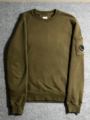🧥 C.P. Company – Olive Lens Sweatshirt (M) - Stylish and iconic C.P. Company sweatshirt in a beautiful olive green tone. Size M, length 67 cm, width 53 cm – fits perfectly for a modern, relaxed look. The sleeve features the classic lens detail, instantly recognizable to fans of the brand.  ✅ Condition: Excellent – no wear, no stains 💯 Authenticity: 100% genuine, you can verify it easily on certilogo.com 📦 Shipping: Dispatched immediately after purchase – fast and hassle-free delivery!  This sweatshirt combines C.P. Company’s technical styl