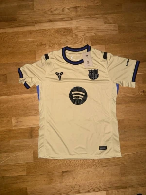 Barcelona bortatröja Raphinha 11 - Snygg beige Barcelona bortatröja från Nike med Raphinha 11 på ryggen. Tröjan har svarta detaljer, klubbmärke och sponsorloggor på bröstet samt Champions League-märke på ärmen. Tillverkad i lätt och ventilerande material, perfekt för fotboll eller träning.