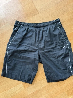 Svarta träningsshorts från Nike - Svarta träningsshorts från Nike med vit piping längs sidorna och elastisk midja. Lätta och luftiga, perfekta för sport eller gym. Klassisk Nike-logga på vänster ben och enkel design som funkar till det mesta.