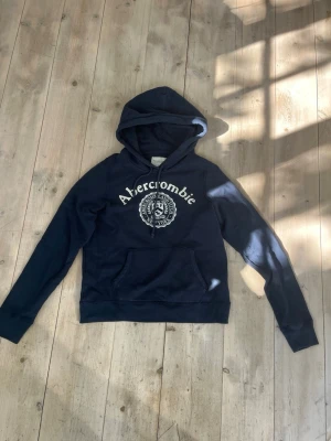Mörkblå hoodie från Abercrombie - Snygg mörkblå hoodie från Abercrombie med vit logga och emblem på bröstet. Klassisk känguruficka framtill och justerbar huva. Tillverkad i mjukt bomullsmaterial som är skönt mot huden. Perfekt för en avslappnad och trendig look.