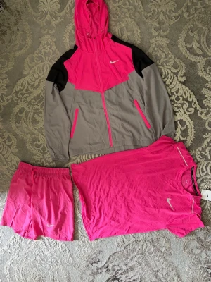 Nike träningsset i rosa och grått - Säljer ett sportigt träningsset från Nike som består av en rosa och grå vindjacka med huva, matchande rosa shorts och en rosa t-shirt. Jackan har dragkedja, svarta axelpartier och två fickor. Perfekt för löpning eller gymmet. Tracksuiten är i storlek S, shortsen i storlek M och t shirten i storlek L