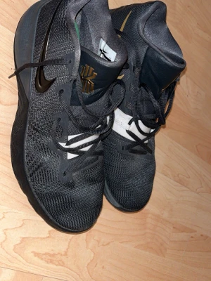 Nike Kyrie svarta basketskor - Snygga svarta Nike Kyrie basketskor med guld- och vitdetaljer. Ovandelen är i mesh för bra andningsförmåga och sulan har gröna, vita och svarta partier med bra grepp. Perfekta för basketplanen och streetstyle.