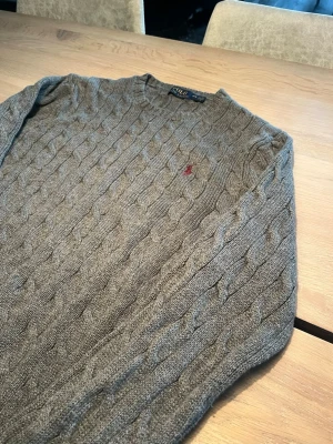 Grå kabelstickad tröja Polo Ralph Lauren - Kabelstickad grå tröja från Polo Ralph Lauren med rund halsringning och klassisk röd broderad logga på bröstet. Tröjan har långa ärmar och är tillverkad i ett mjukt, värmande material som passar perfekt till kyligare dagar.