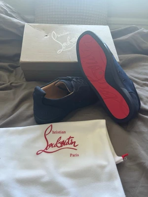 Louboutins skor - Snygga svarta sneakers med ikonisk röd sula och diskret logga. Skorna har lågt skaft och är tillverkade i mocka och läder. Perfekta för dig som vill sticka ut med exklusiv design och streetkänsla.