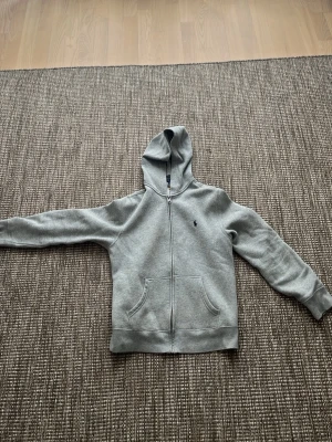 Grå hoodie från Polo Ralph Lauren - Snygg grå hoodie från Polo Ralph Lauren med dragkedja framtill och klassisk huva. Har två stora fickor på magen och den ikoniska lilla loggan broderad på bröstet. Perfekt för en chill och stilren look.