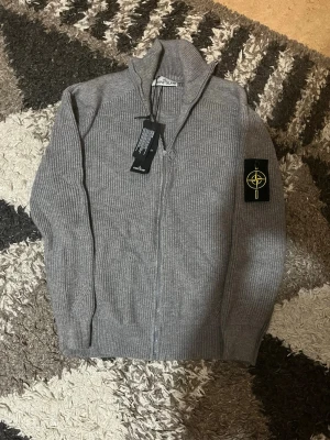 Grå ziptröja från Stone Island - Snygg grå ribbstickad ziptröja från Stone Island med hög krage och ikonisk logotyp på vänster ärm. Tröjan har hel dragkedja framtill och är tillverkad i mjukt stickat material. Perfekt för lager-på-lager och streetwear-stil.