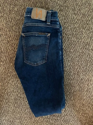 Mörkblå jeans från Nudie Jeans - Säljer ett par klassiska mörkblå jeans från Nudie Jeans med snygga kontrastsömmar och bakficka med broderad detalj. Jeansen har en rak passform och är tillverkade i slitstark denim. Perfekta för dig som gillar stilrena och tidlösa jeans.