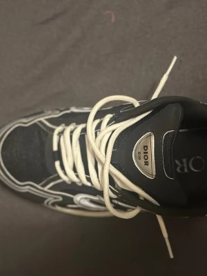 Dior B30 svarta sneakers med vita detaljer - Snygga Dior B30 sneakers i svart med vita kontrastdetaljer och vita skosnören. Skon har en chunky sula och stora Dior-loggor på sidan. Ovandelen är i mesh och läder för en modern och sportig vibe.